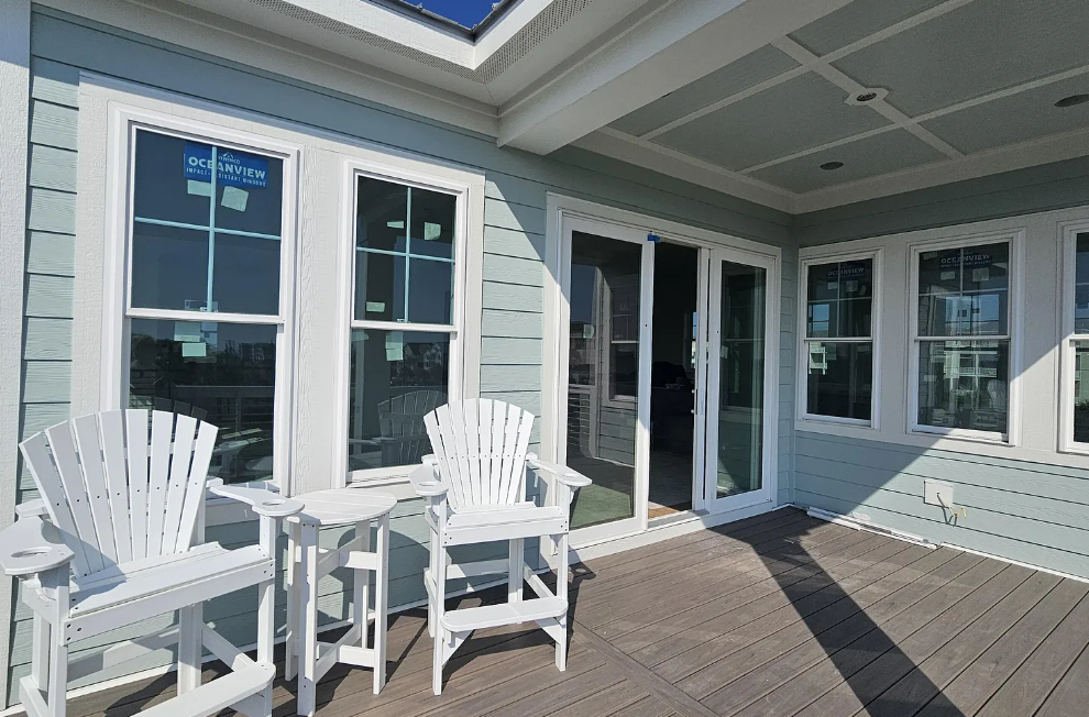 Patio with white chairs and small table; sliding glass doors, light blue siding, and wood decking.
