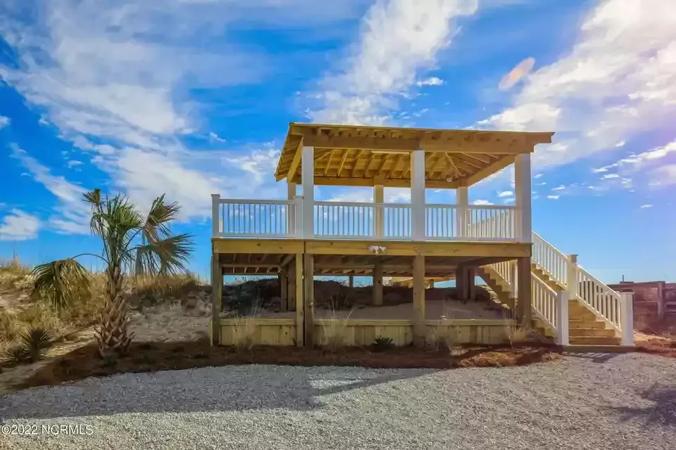 A house with a large deck and stairs is sitting on top of a gravel lot.