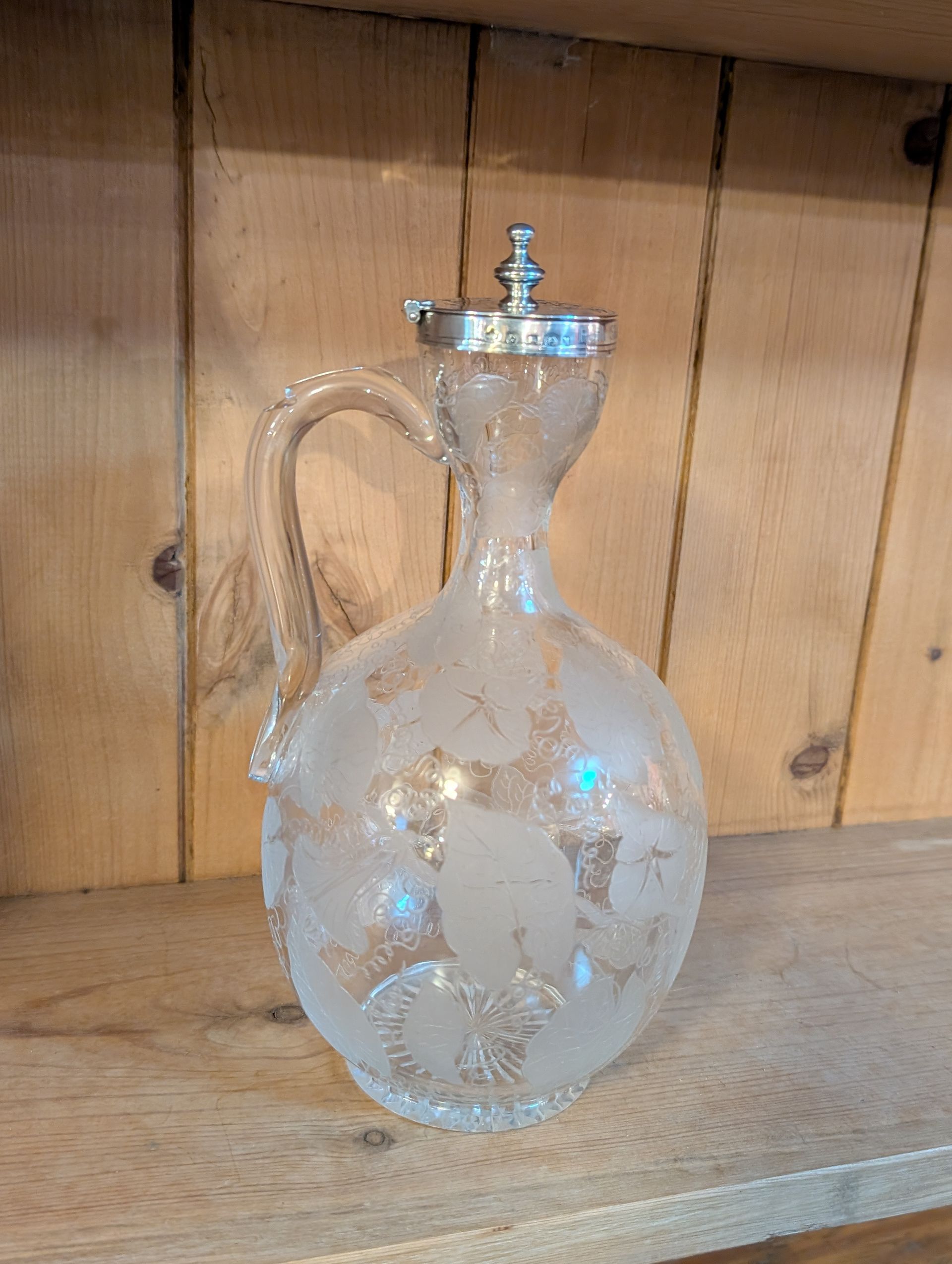 A clear glass pitcher with a handle is sitting on a wooden shelf.