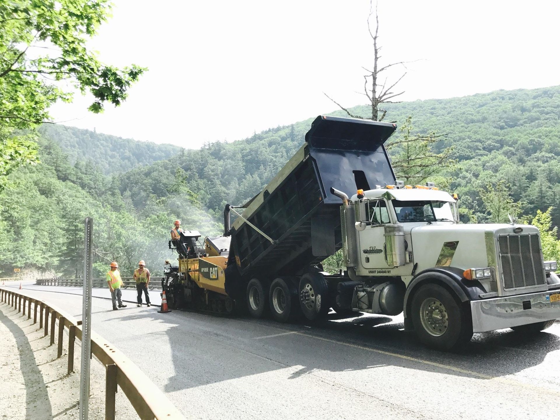 Dump truck unloading asphalt into a paver on a mountain road, workers nearby.