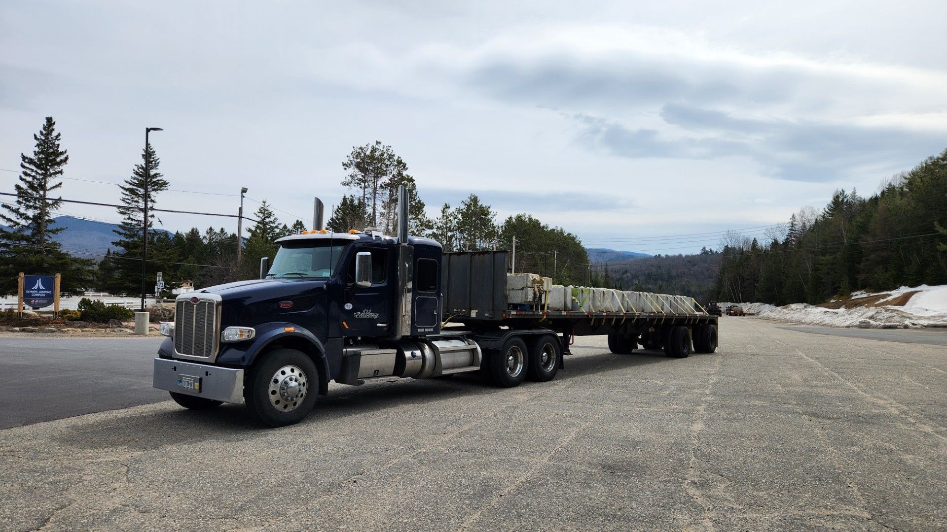 Black semi-truck with trailer parked on a gravel lot near trees under cloudy sky