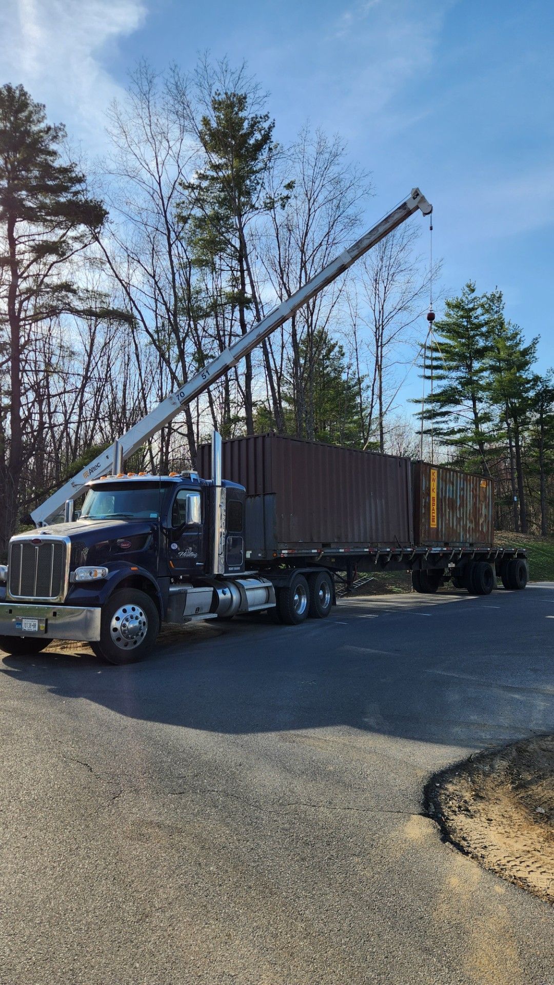 Semi truck with raised crane arm hauling a large container on a paved road in a wooded area