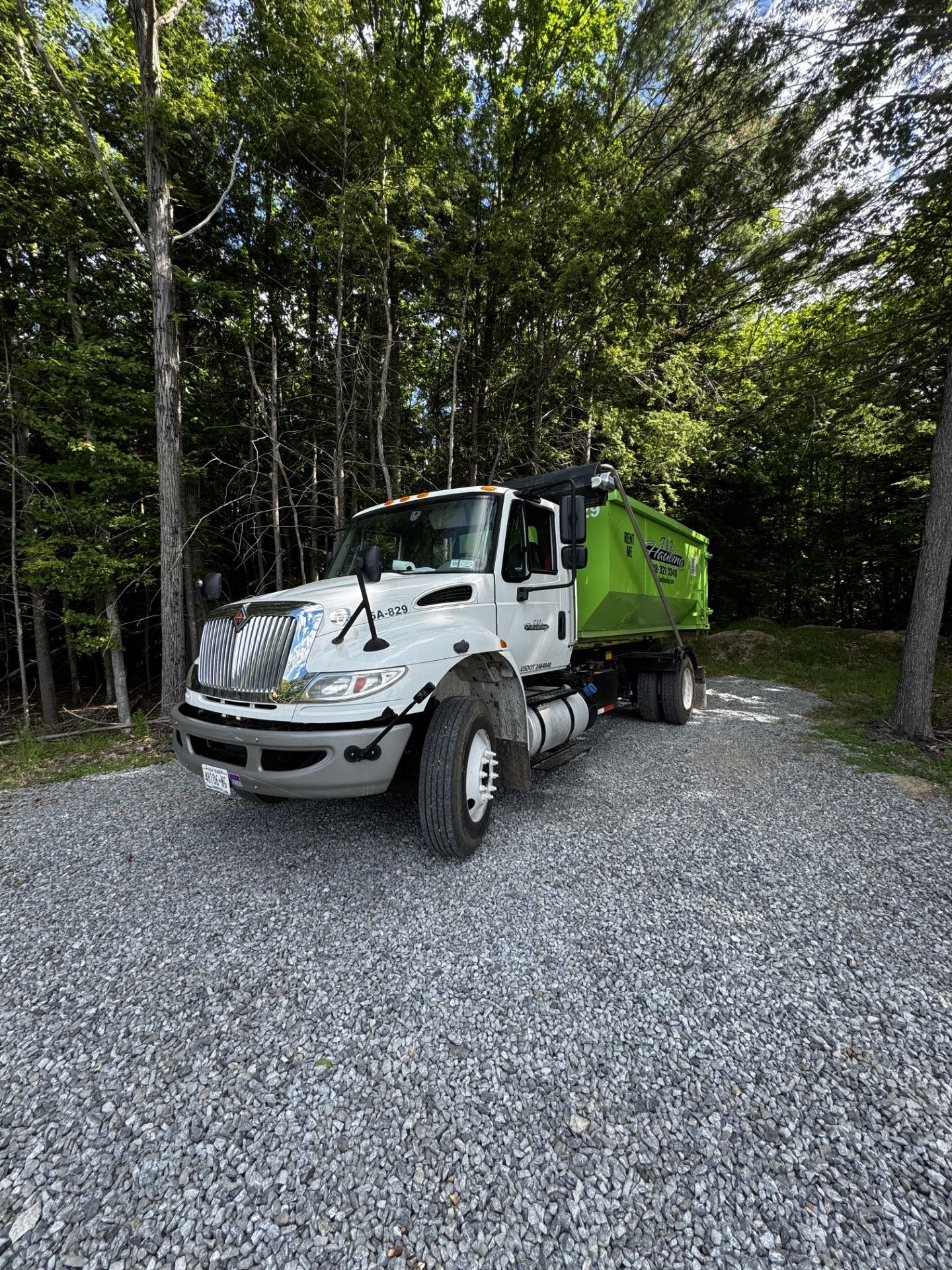 White and green dump truck parked on gravel beside trees in a wooded area