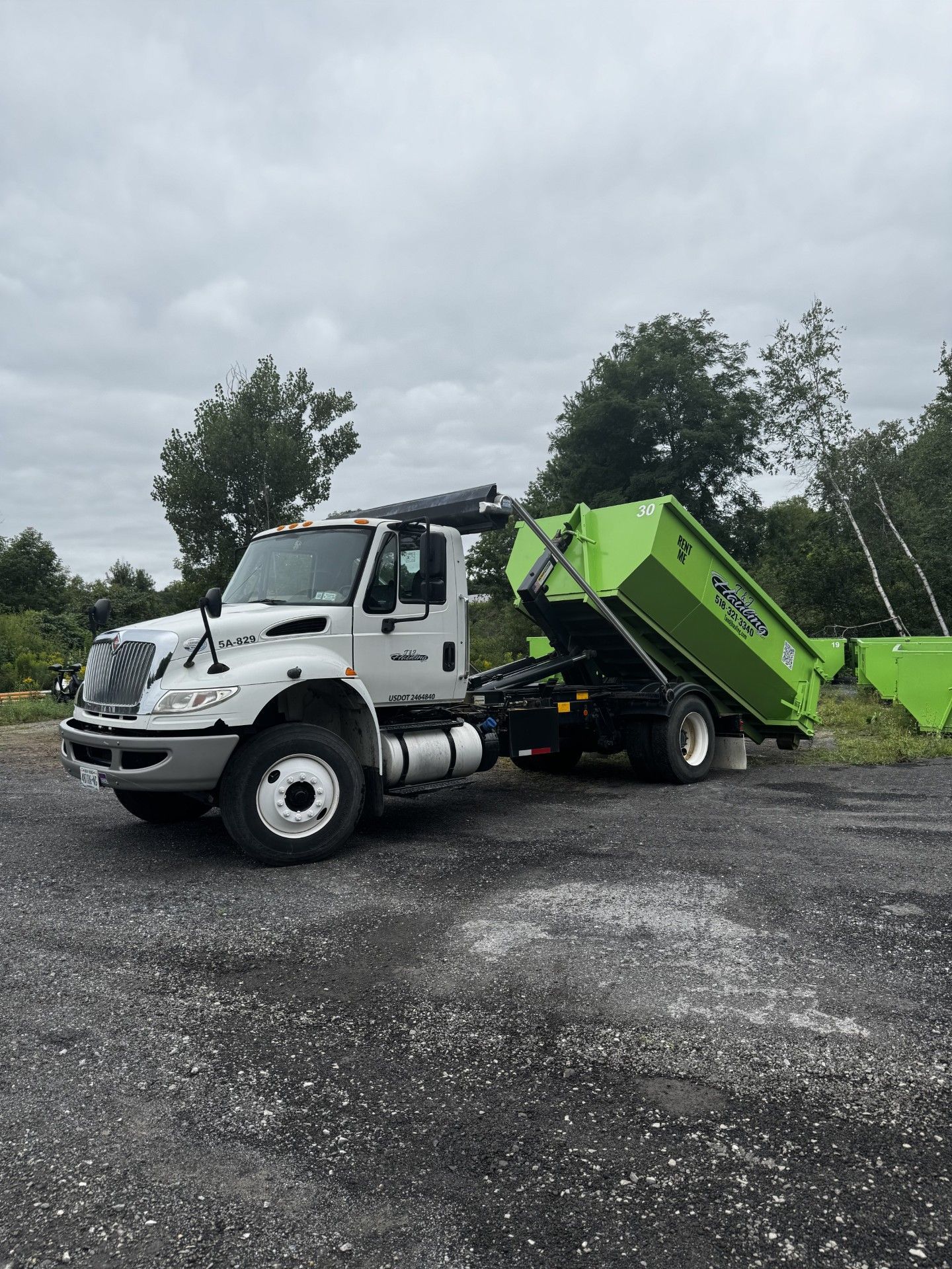 Green dump truck parked on gravel under cloudy sky, rear bed raised at a dump site