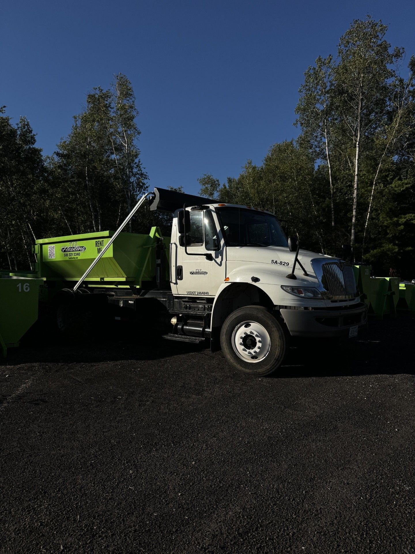 White dump truck with green bed parked on a gravel lot by trees under a blue sky