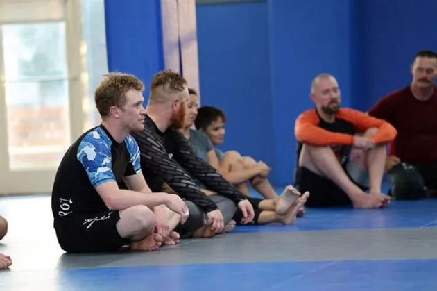 People sitting cross-legged on blue mats in a martial arts class, listening attentively