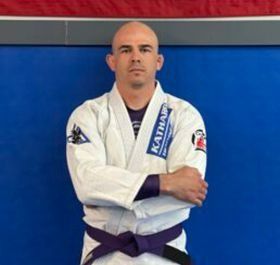 Martial artist in a white gi with a purple belt, standing with arms crossed against a blue mat wall.