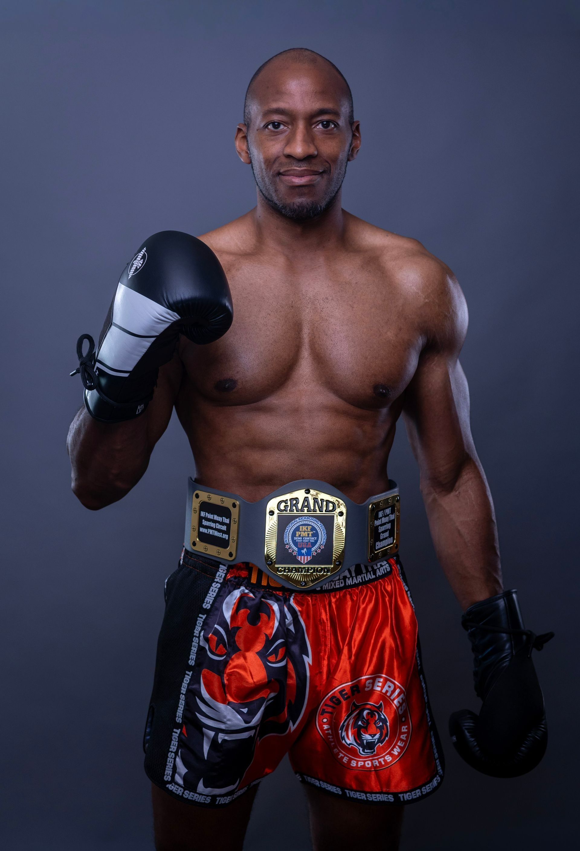 Boxer posing in black gloves and red trunks with a championship belt against a gray backdrop