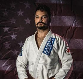 Man in a white martial arts gi with blue patches, standing before a faded American flag backdrop.