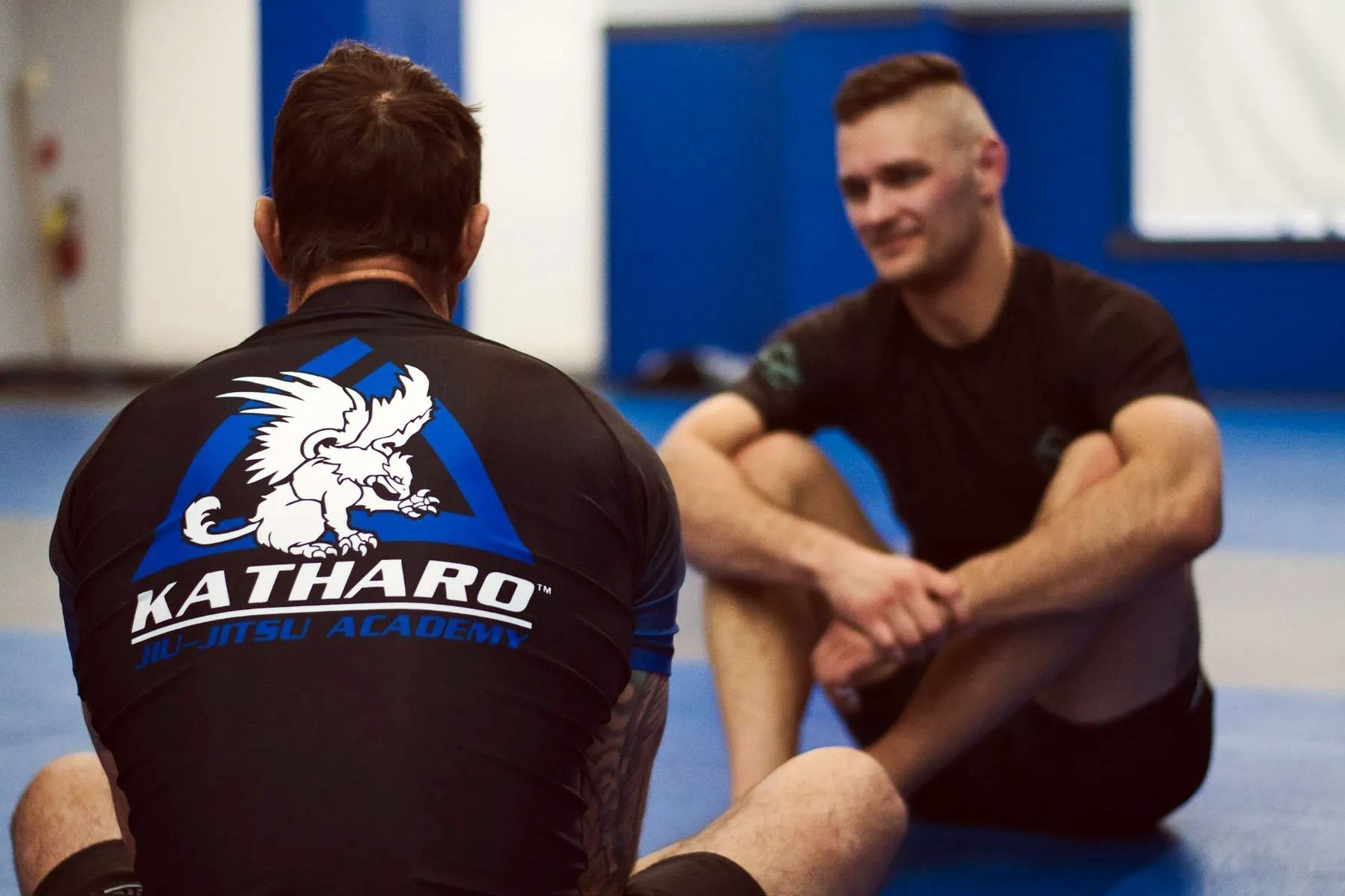 Two men stretching on gym mats, one in a black HATHA logo shirt, in a training studio.