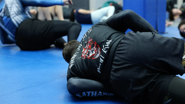Two grapplers in black uniforms wrestling on a blue mat in a gym