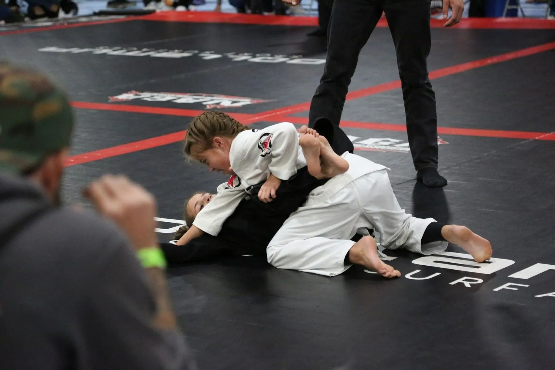 Two grapplers in white gis on a black mat during a jiu-jitsu match, with a referee standing nearby