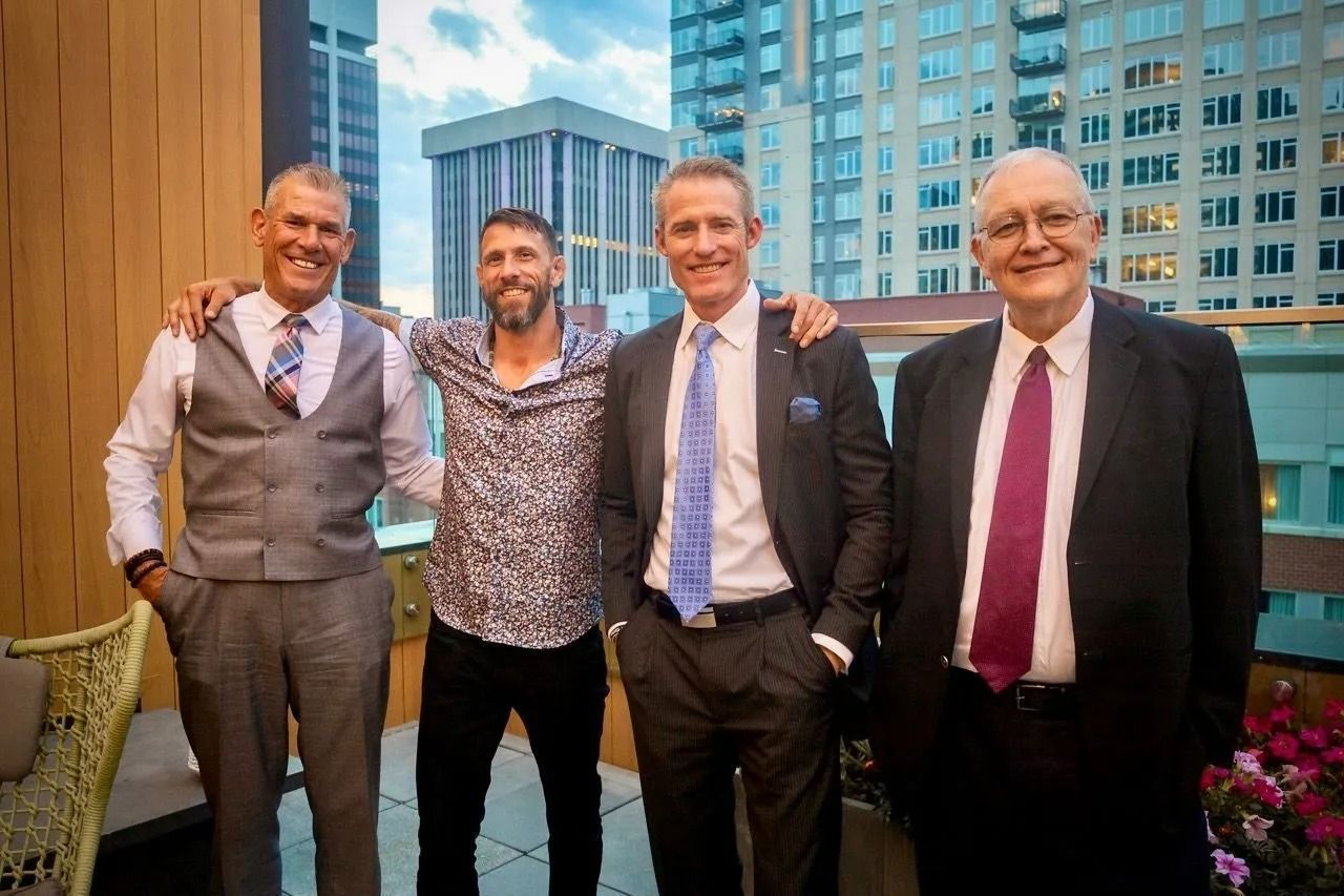 Four men standing together outdoors on a city rooftop, smiling in formal attire.