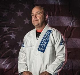 Man in a white martial arts gi with a blue belt, standing before a faded American flag backdrop.