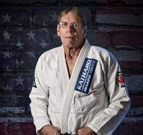 Person in a white martial arts gi with a navy name patch, standing against a dark American flag background.