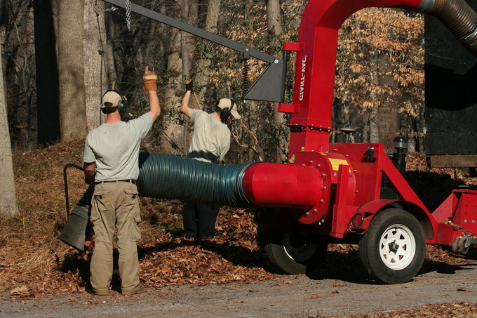Two workers loading leaves into a red wood chipper beside a tree-lined road.