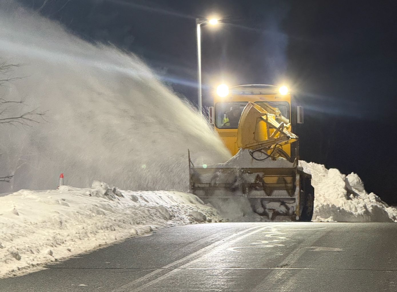 Snowplow clearing heavy snow on a road at night under streetlights.