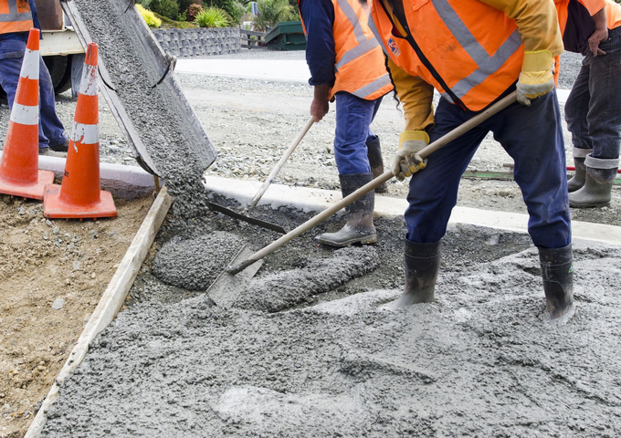 Workers in orange vests pouring wet concrete on a road beside traffic cones.