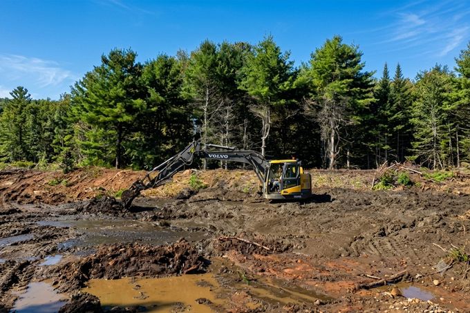 Excavator working at a muddy construction site with a dirt mound and forest backdrop