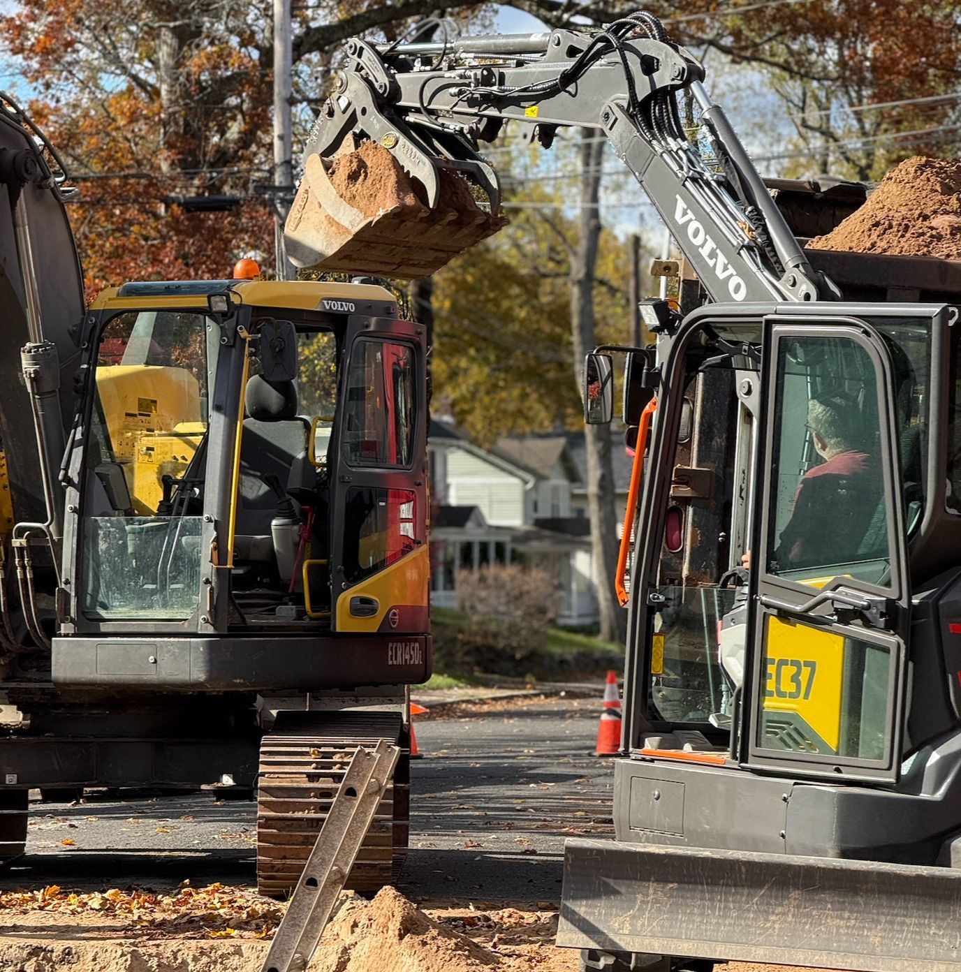 Two excavators load debris into a truck on a street lined with autumn trees.
