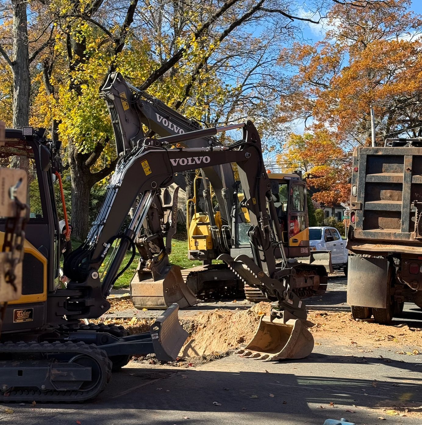 Volvo excavator at a construction site beside trees with autumn leaves, surrounded by heavy equipment.