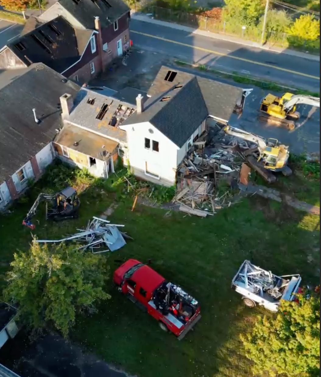 Aerial view of storm-damaged homes with debris, a red pickup, and a yellow excavator by a road.