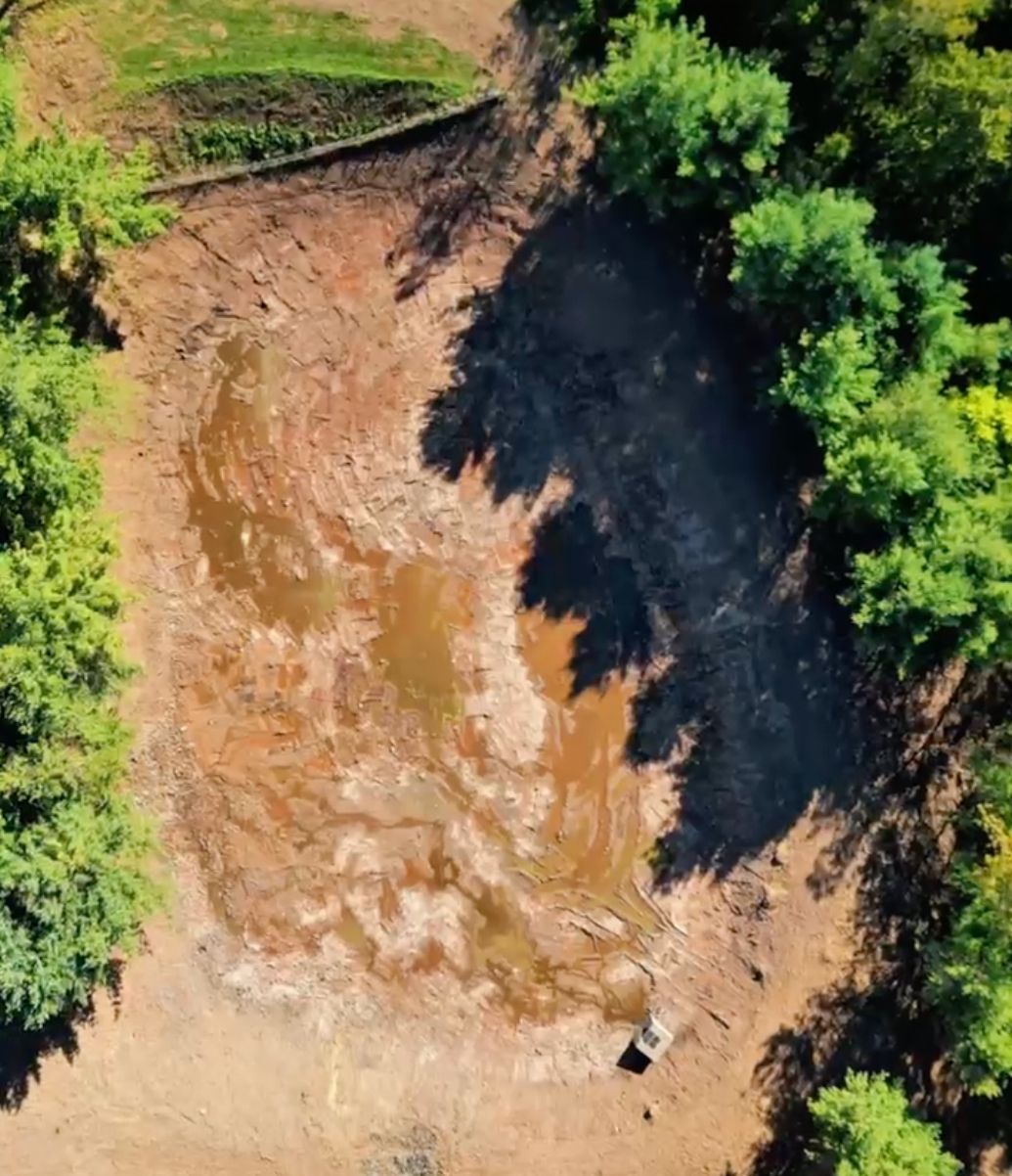 Aerial view of a muddy cleared area bordered by dense green trees and dark shadow.