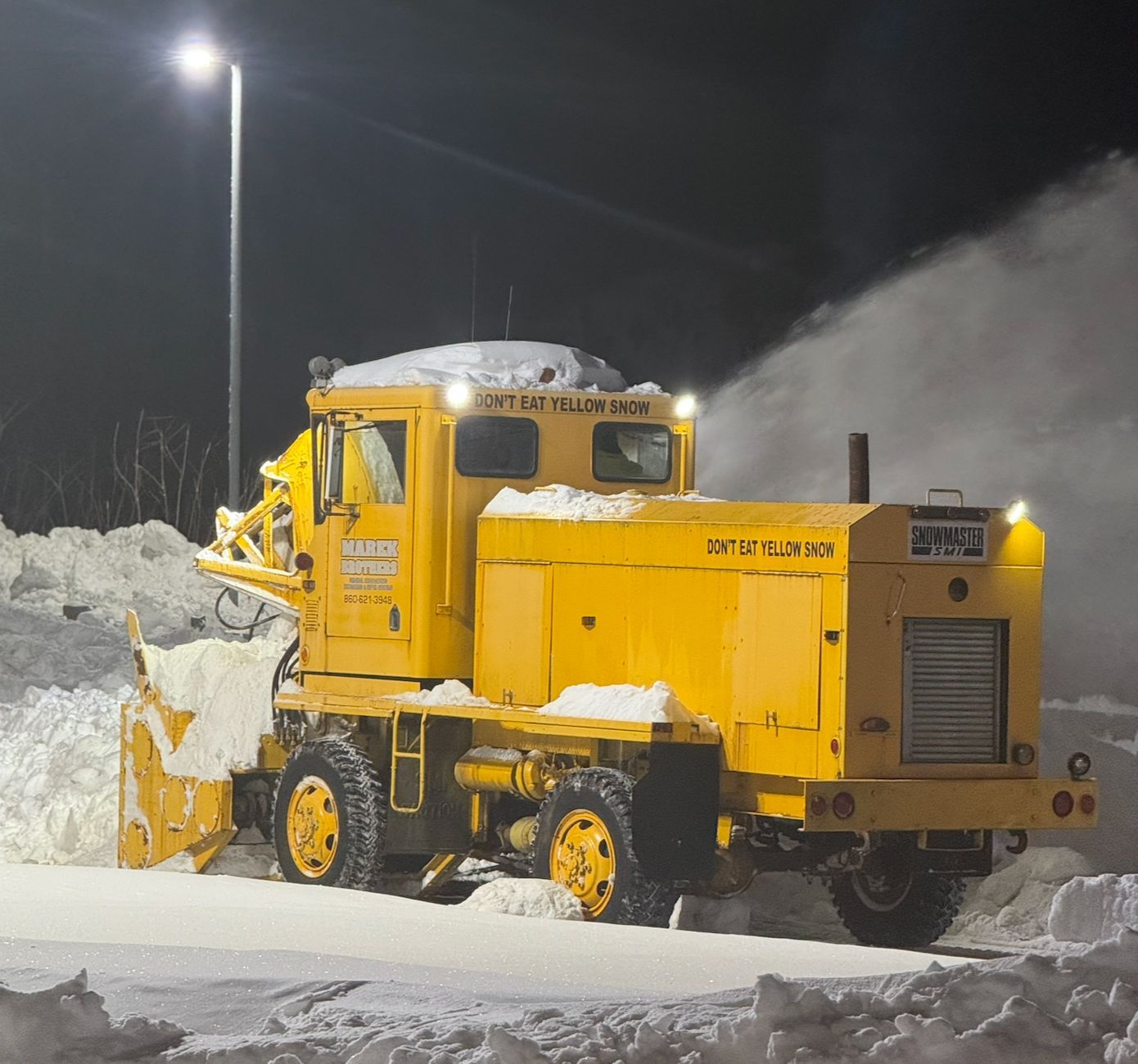 Yellow snowplow truck clearing snow at night under streetlights.