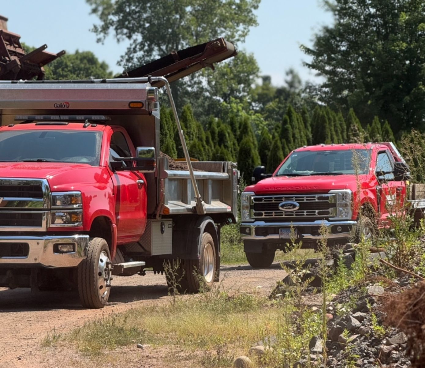 Two red pickup trucks on a gravel path, one loaded with equipment in a wooded area.