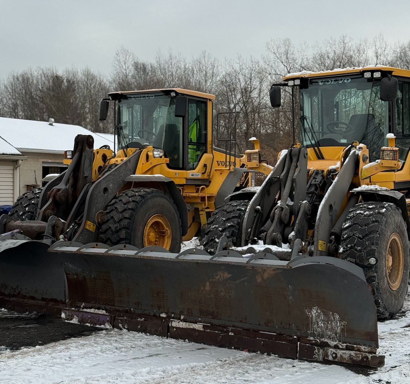 Two yellow front-end loaders with a snowplow blade parked in a snowy lot