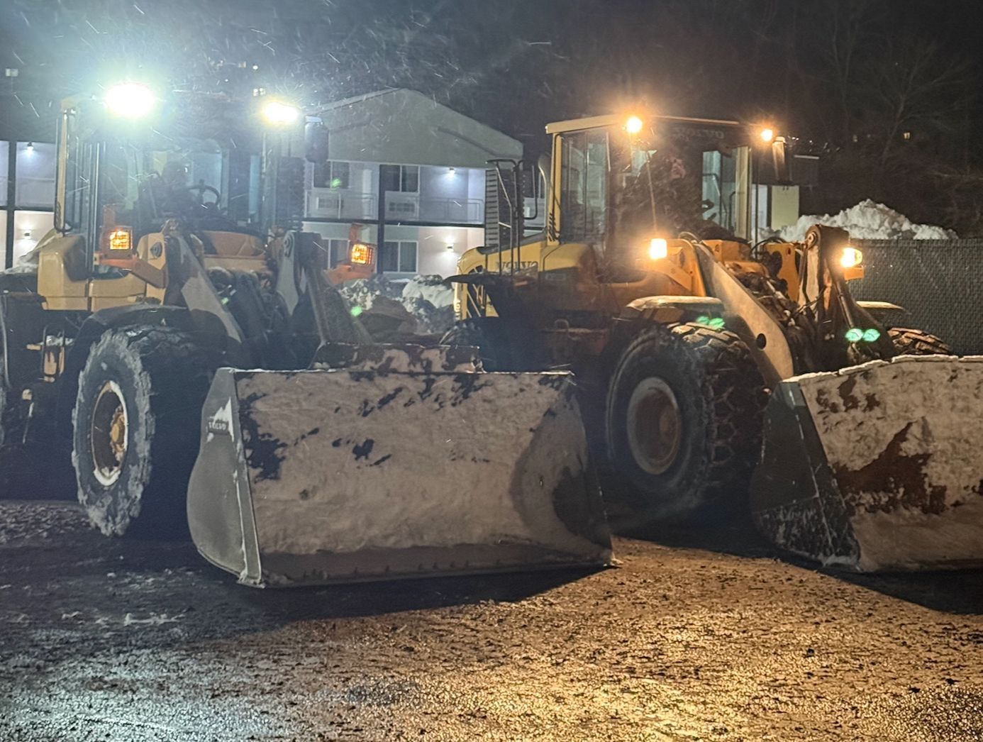 Two large front loaders parked at night on a wet street, lights reflecting on the ground.