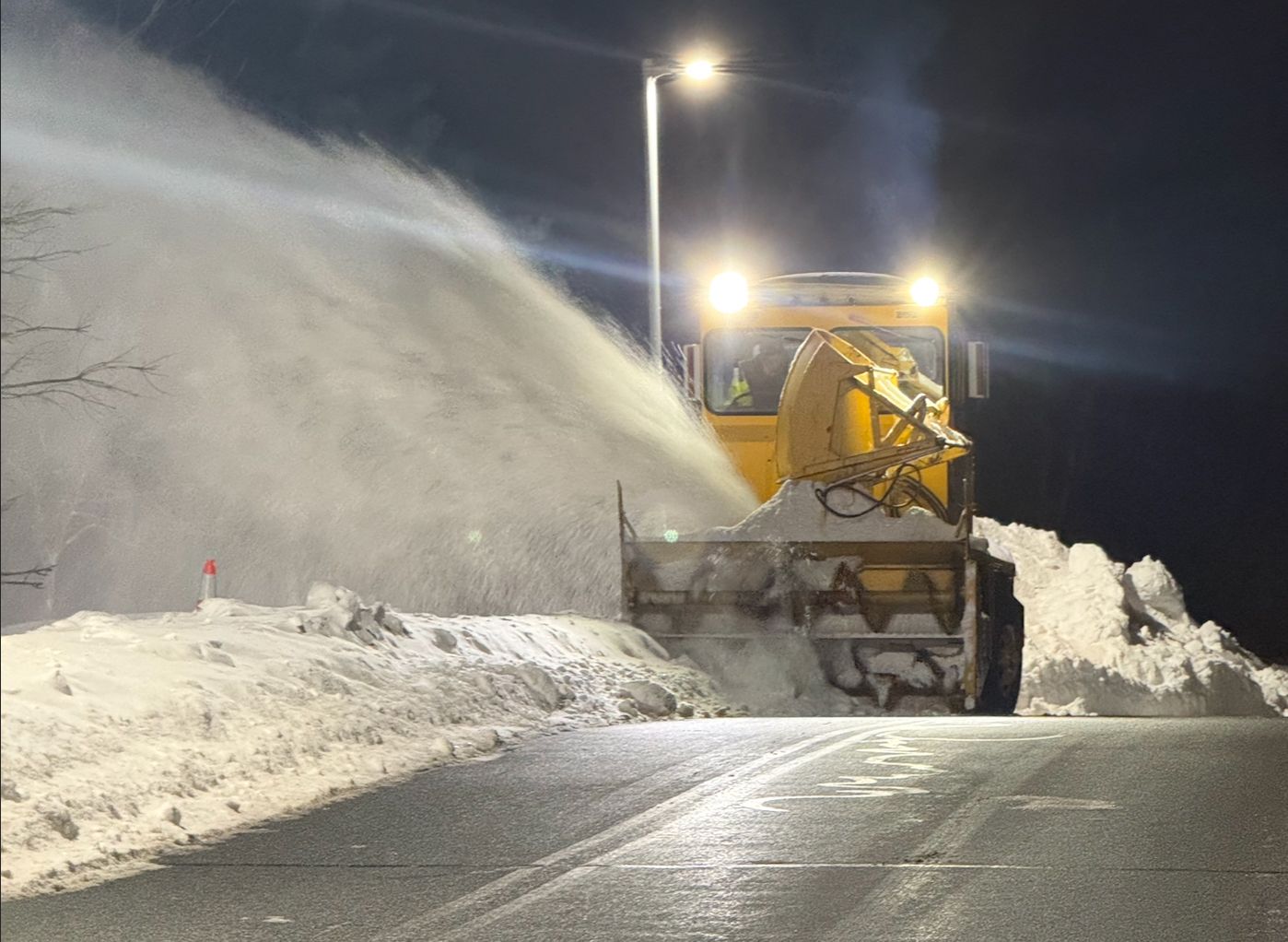Yellow snowplow clearing a road at night, spraying snow under bright streetlights.