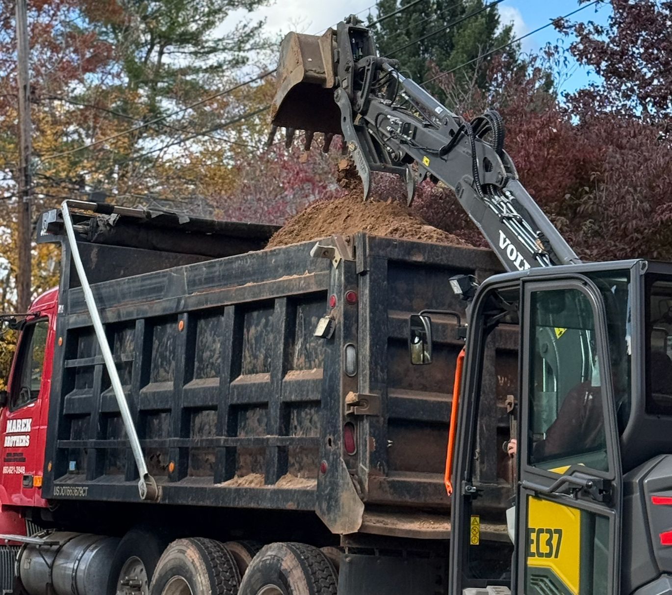 Excavator loading dirt into a black dump truck outdoors in autumn.