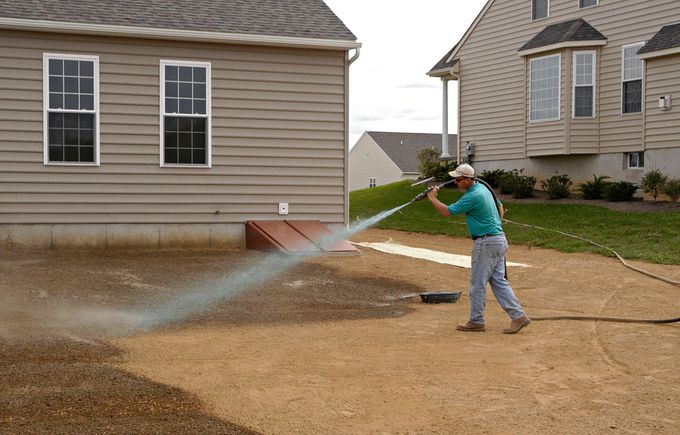 Person spraying water from a hose across a dirt yard beside suburban houses.