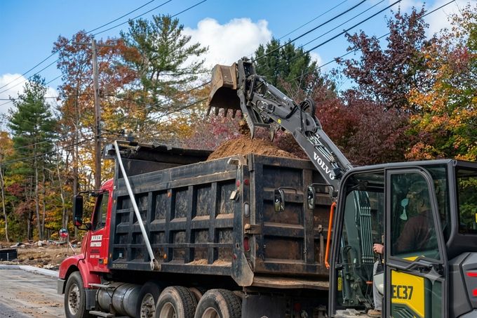 Excavator loading debris into a black dump truck on a roadside with autumn trees.