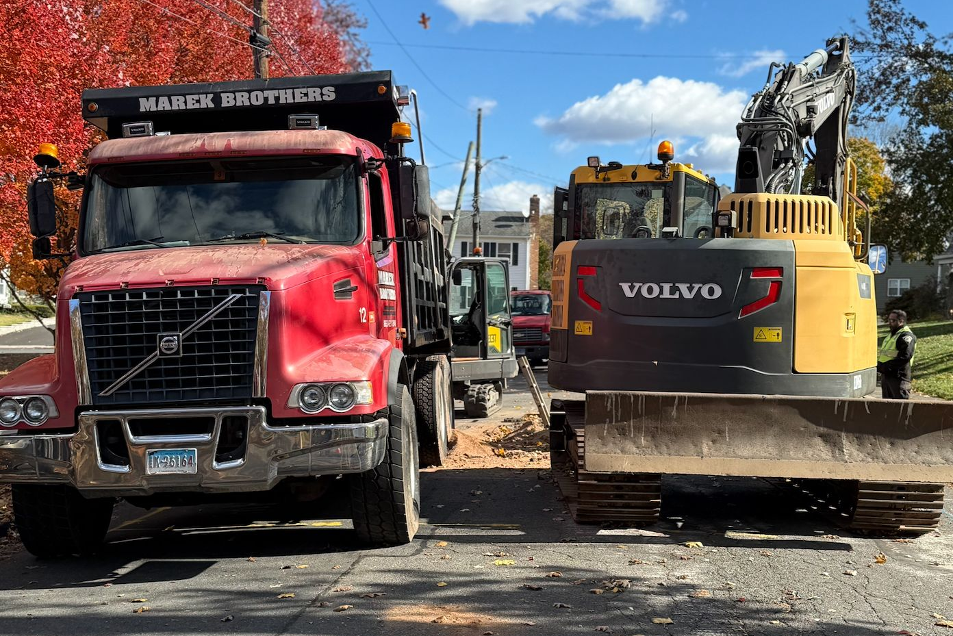 Red Volvo truck and yellow excavator parked side by side on a dirt road under a blue sky.