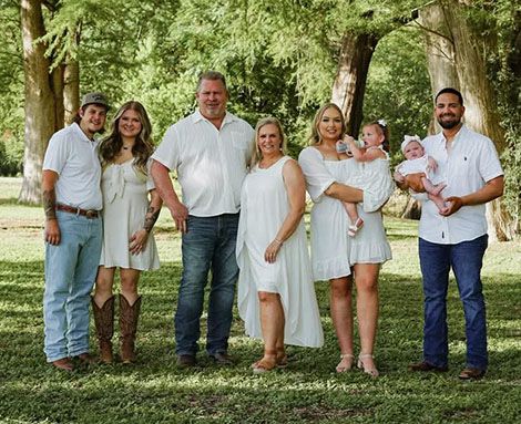 Family of 8 in white outfits and jeans poses outdoors on grass in front of trees.