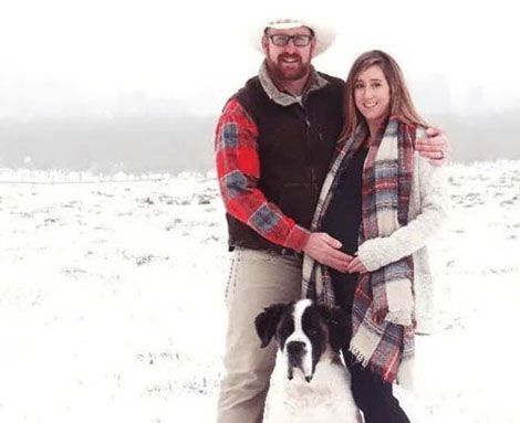 A man and a pregnant woman are standing in the snow with a dog.