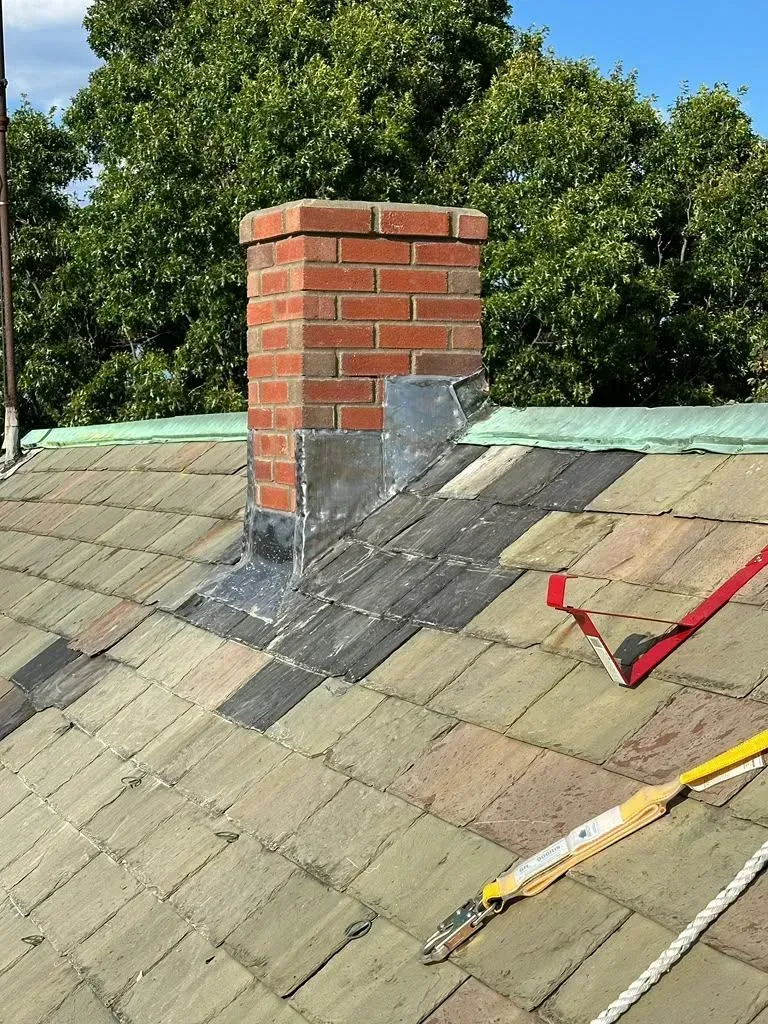 Brick chimney on a tiled roof with metal flashing and tools nearby under trees