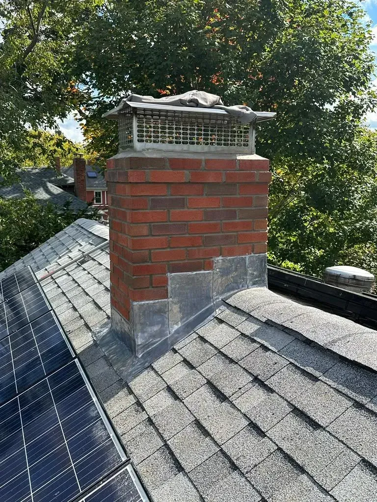 Brick chimney on a gray shingled roof with solar panels, surrounded by trees.