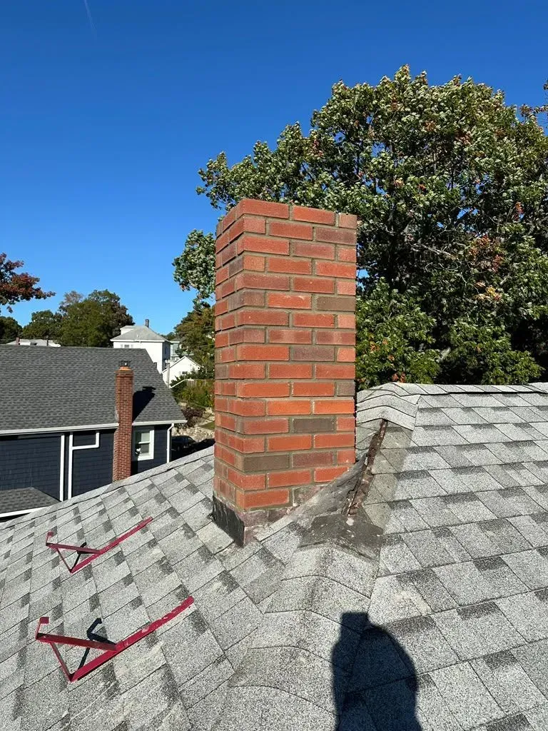 Brick chimney on a shingled rooftop under a clear blue sky, with trees and neighboring houses in the background.