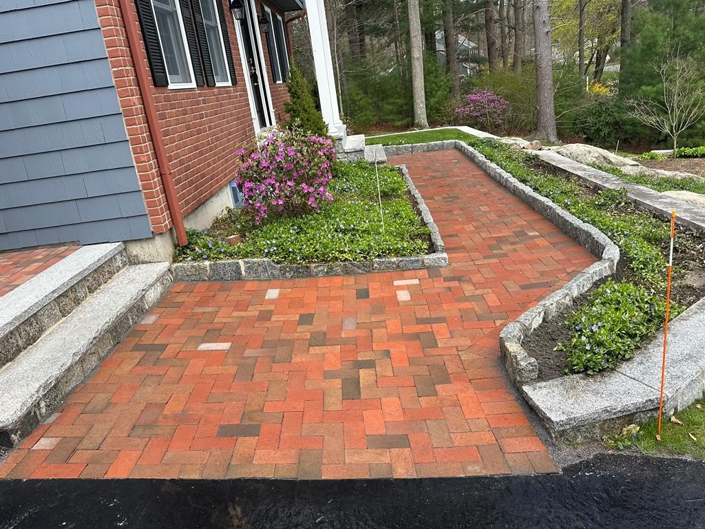 Brick walkway beside a house, bordered by garden beds and a small flowering shrub.