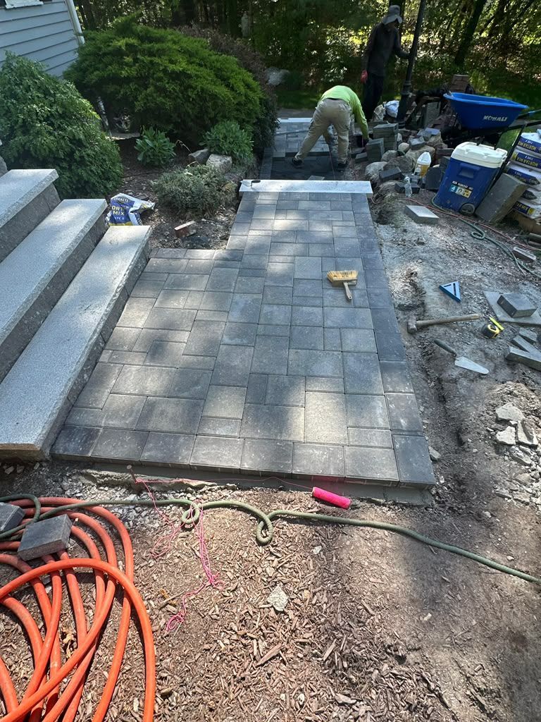Brick paver walkway under construction beside garden steps, with workers and tools visible.