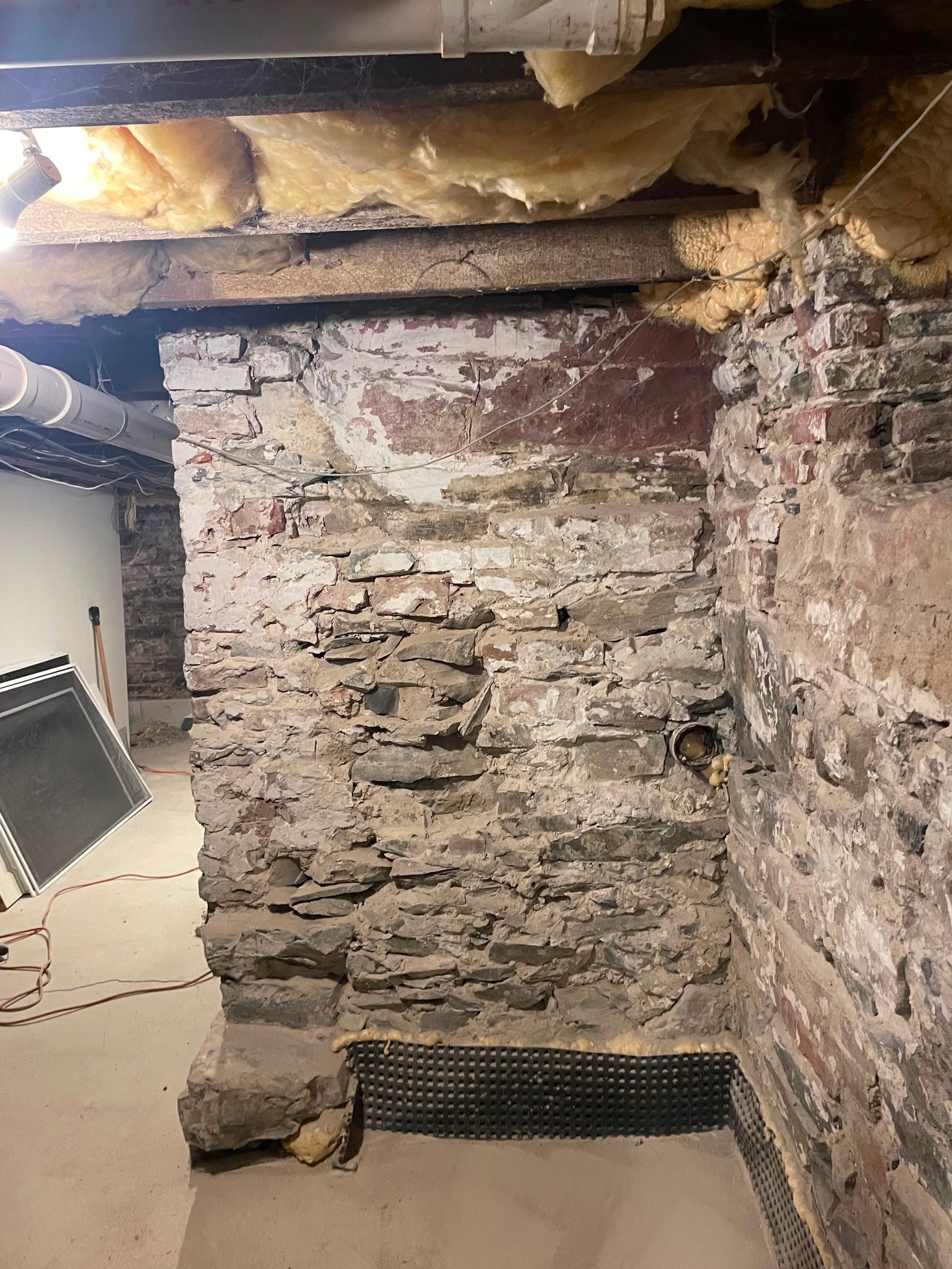 Damp stone basement wall with peeling paint, exposed beams, and yellow insulation overhead.