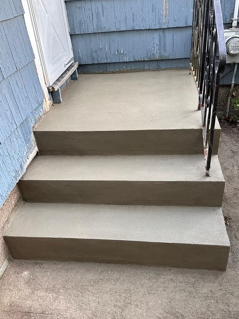 Three concrete steps leading to a blue-sided house entrance with a black metal railing.