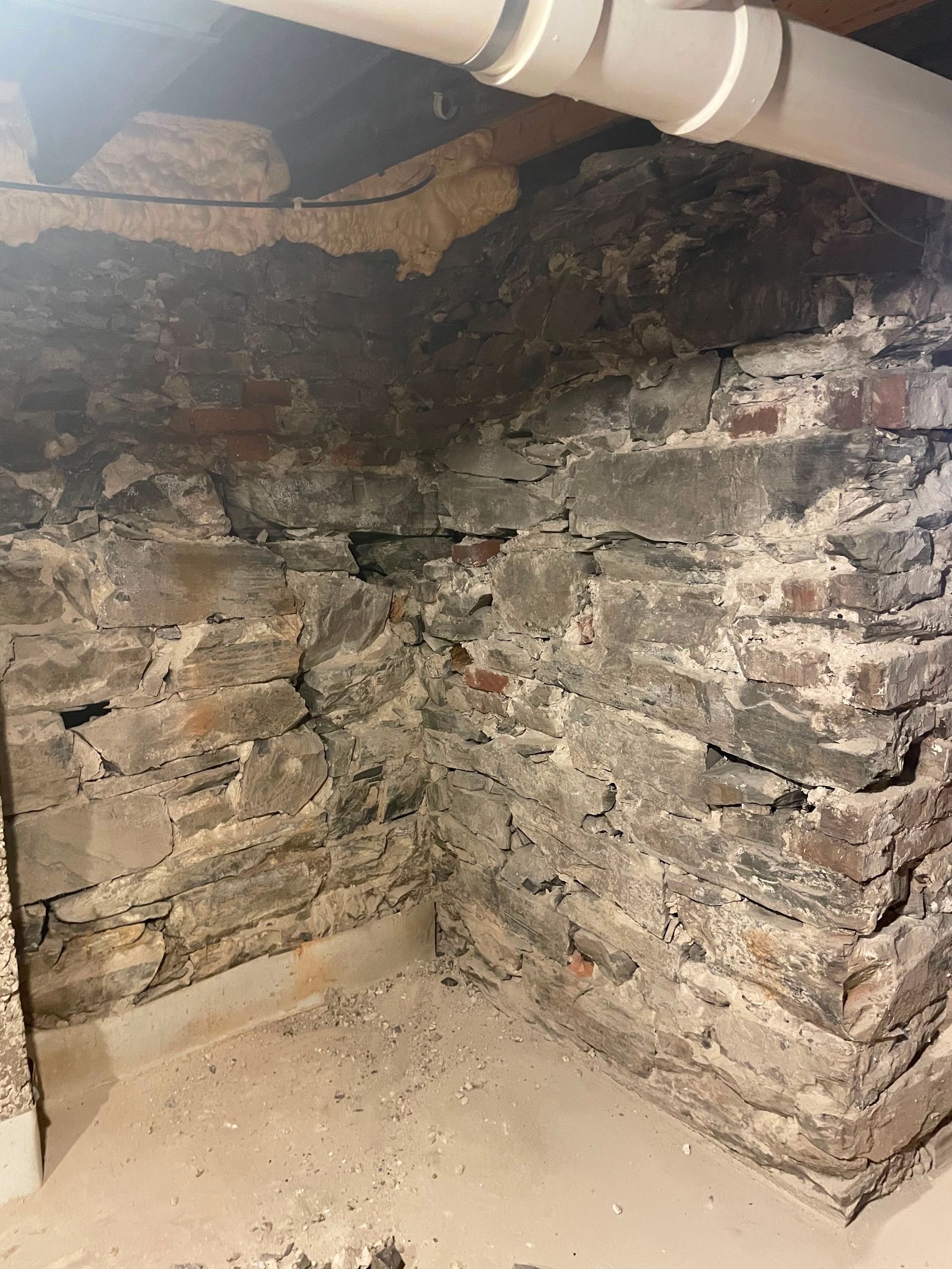 Damp basement corner with crumbling stone foundation, exposed pipe, and peeling plaster.