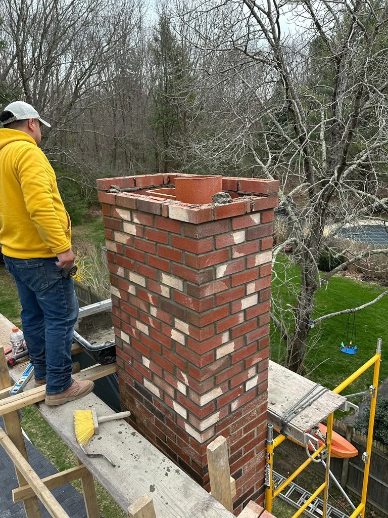 Brick chimney under construction on a rooftop, with a worker in a yellow jacket nearby.