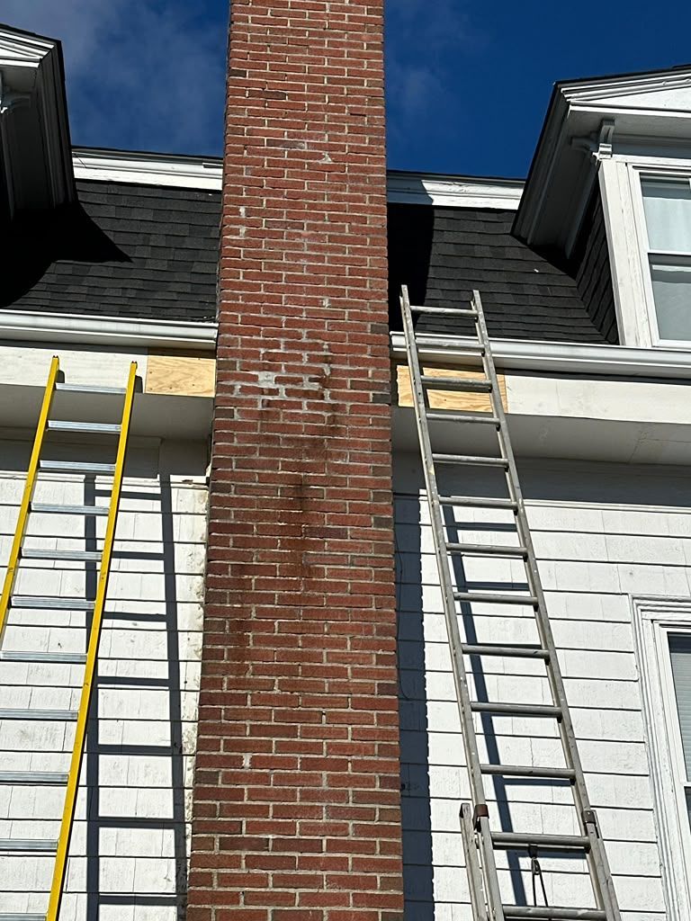 Ladders leaning against a brick chimney on a white house with gray roof shingles