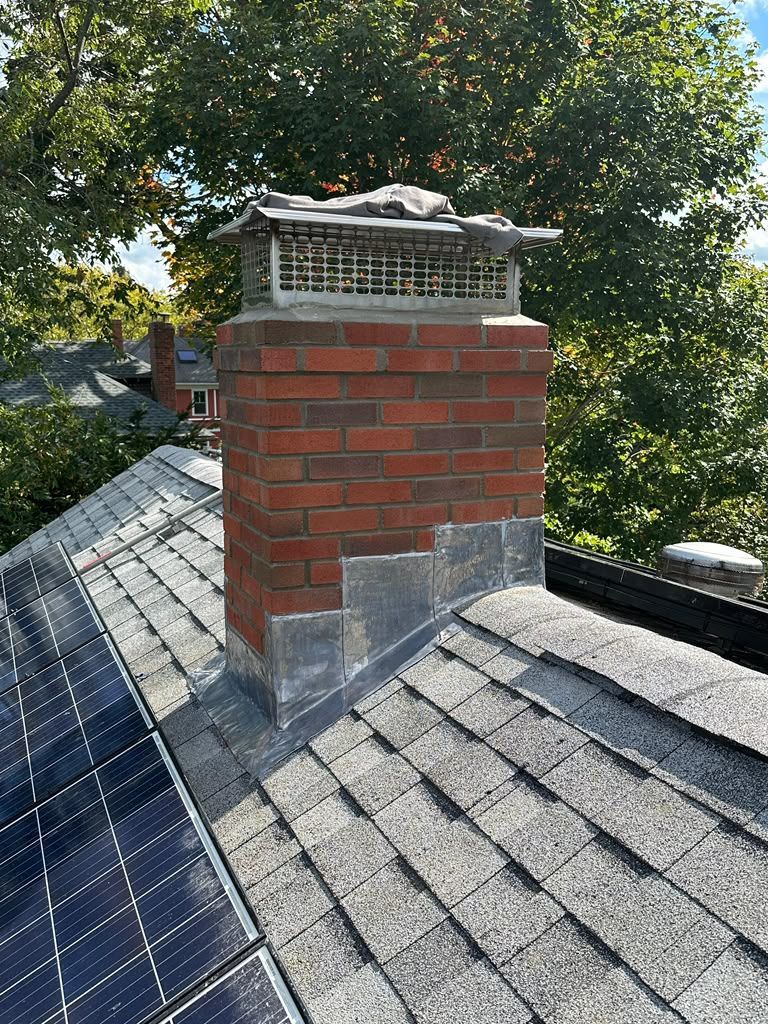Brick chimney with metal cap on a shingled roof beside solar panels and trees
