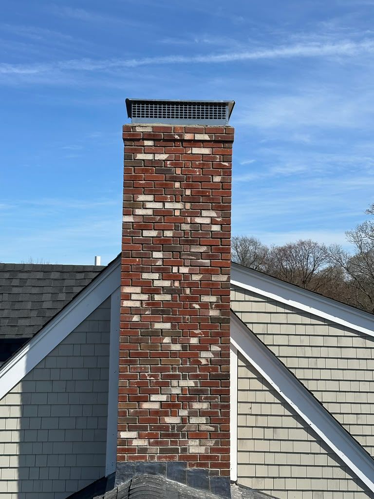 Brick chimney rising above a gray shingled roof under a blue sky.